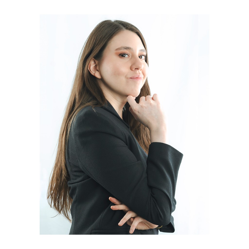 Liz Stryke talent portrait: structured black blazer, dynamic pose with hand at chin and playful smirk; warm makeup on white studio backdrop — actor and model photography.