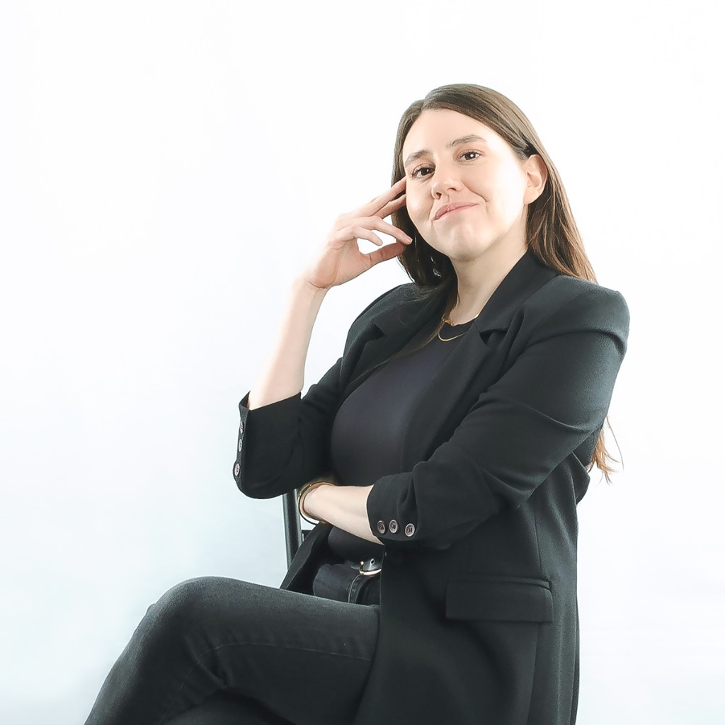 Liz Stryke seated portrait: black blazer, hand at cheek and calm expression; gold necklace and bangle visible; soft studio backdrop — natural corporate portrait.