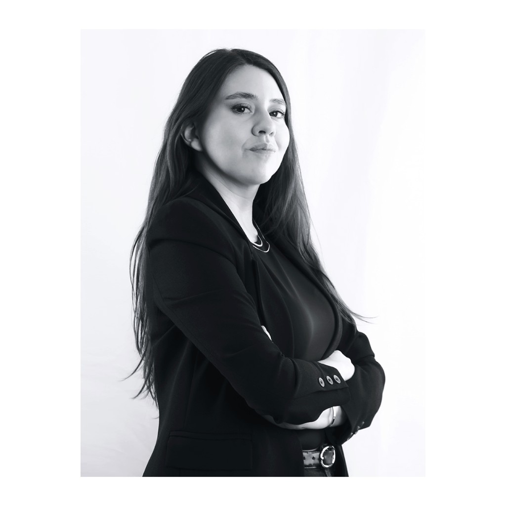 Liz Stryke B&W profile portrait: black blazer, arms crossed and belt, three-quarter over-shoulder look; side light on white backdrop — assertive talent headshot.