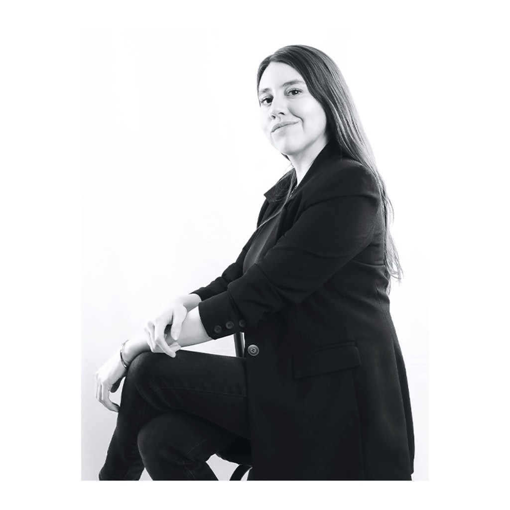 Liz Stryke B&W seated portrait: oversized black blazer, crossed leg and hands on knee; soft smile facing camera on high-key white background — studio talent session.