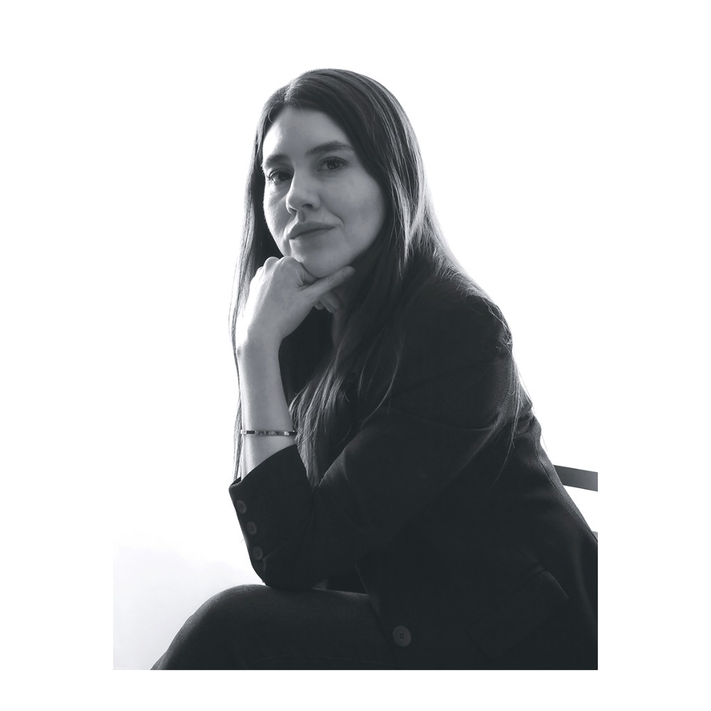 Liz Stryke B&W thoughtful portrait: black blazer, chin resting on fist and direct gaze to camera; fine silver bracelet; high-key white backdrop — editorial studio portrait.