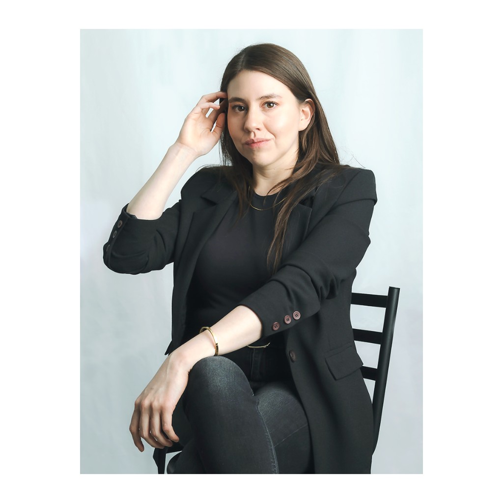 Liz Stryke color portrait: oversized black blazer, dark jeans and black chair, hand at temple and direct gaze; gold bangle on grey-blue studio backdrop — professional talent headshot.