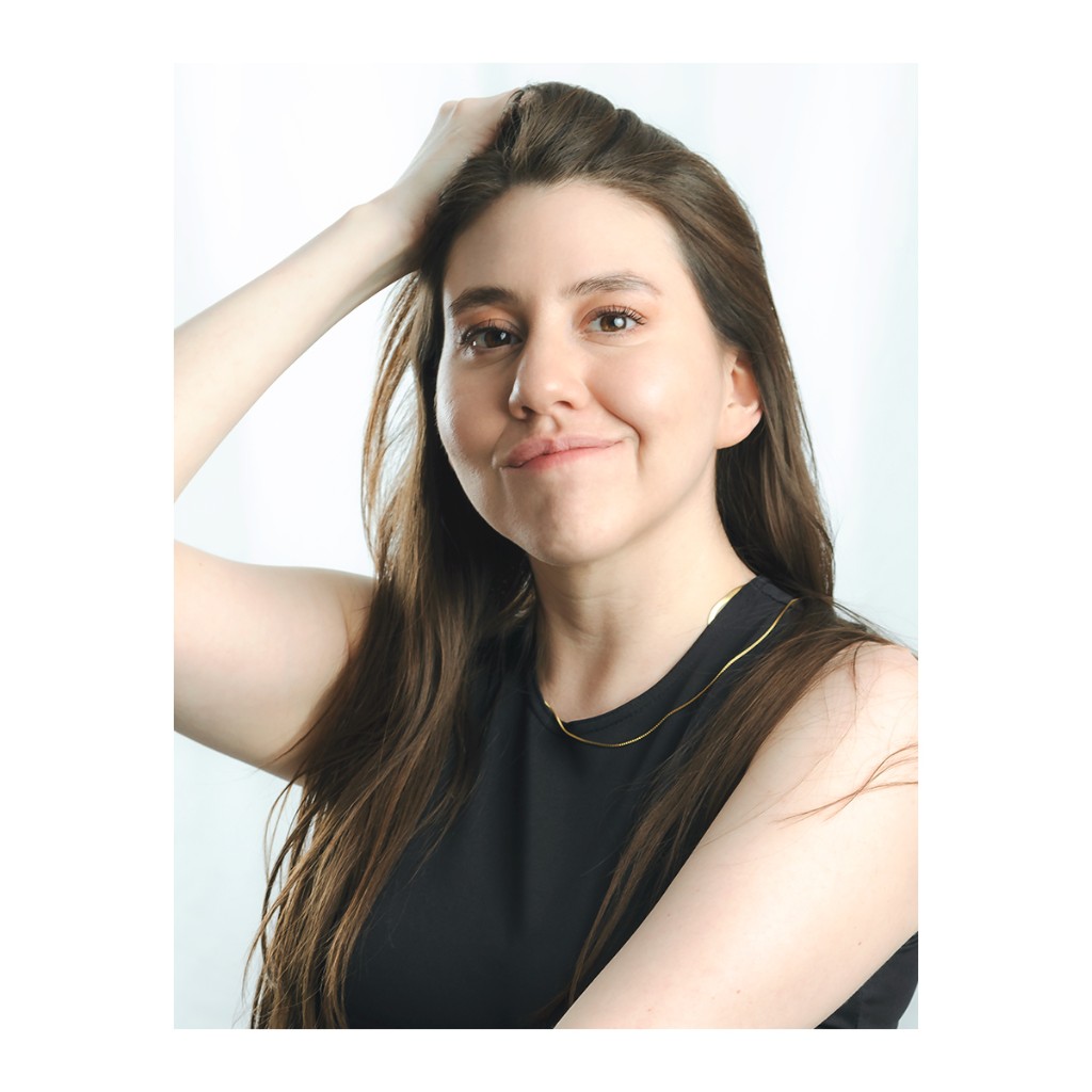 Liz Stryke close-up headshot: black sleeveless tank, gold chain necklace, hand in hair and wide genuine smile; warm makeup on white backdrop — natural talent photography.