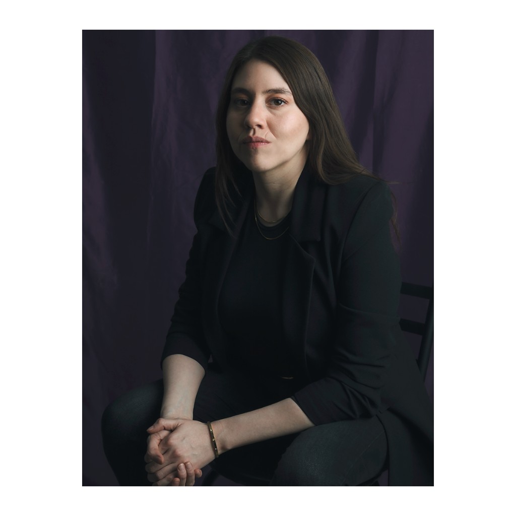 Liz Stryke intimate portrait: dark blazer, hands clasped on lap and gold bangle bracelet; direct pensive gaze against purple curtain — dramatic talent portrait session.