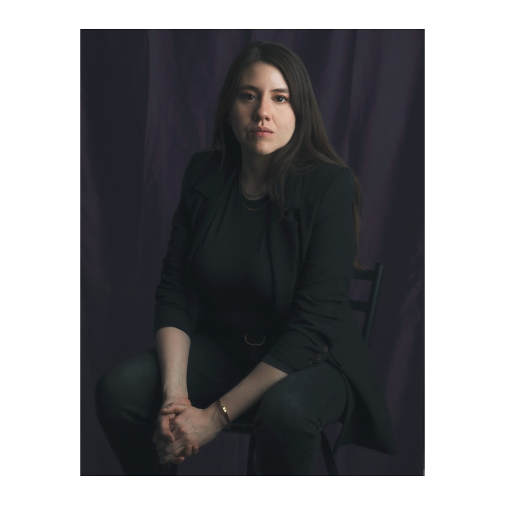 Liz Stryke dramatic portrait: black blazer, dark jeans and O-ring belt, hands clasped; intense gaze to camera against purple curtain backdrop — commercial studio portrait.