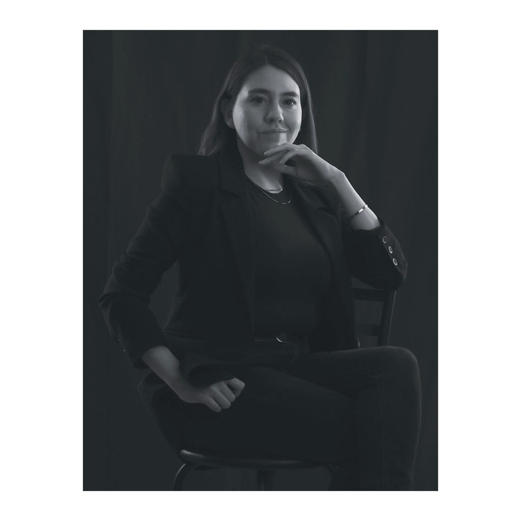 Liz Stryke B&W low-key portrait: black blazer with silver buttons, black belt and seated pose; hand under chin and faint smile; dramatic lighting on dark background — editorial headshot.