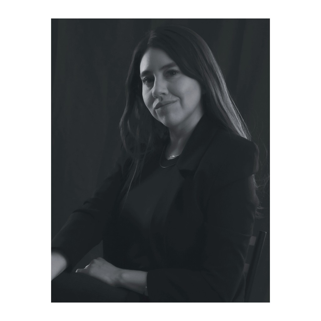 Liz Stryke B&W three-quarter portrait: black blazer, arms crossed in profile pose; warm gaze and soft smile on dark charcoal background — studio talent headshot.