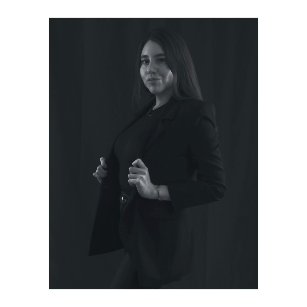 Liz Stryke B&W standing portrait: black blazer with hands on lapels, belt and bracelet; knowing three-quarter look on near-black background — dramatic talent photography.