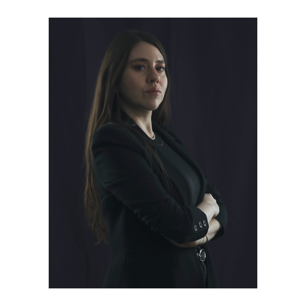 Liz Stryke low-key color portrait: black blazer, dark jeans and O-ring belt, arms crossed; direct confident gaze on deep navy-dark background — dramatic corporate portrait.