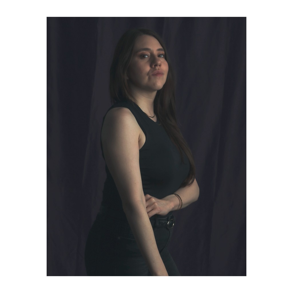 Liz Stryke talent portrait: black sleeveless tank, dark trousers and ring-buckle belt, arms slightly crossed; confident three-quarter profile against dark grey-blue curtain — casual headshot.