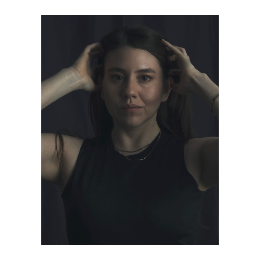Liz Stryke dynamic editorial portrait: black sleeveless tank, both hands raised in hair with a direct gaze to camera; gold chain necklace on dark charcoal background — expressive talent photography.