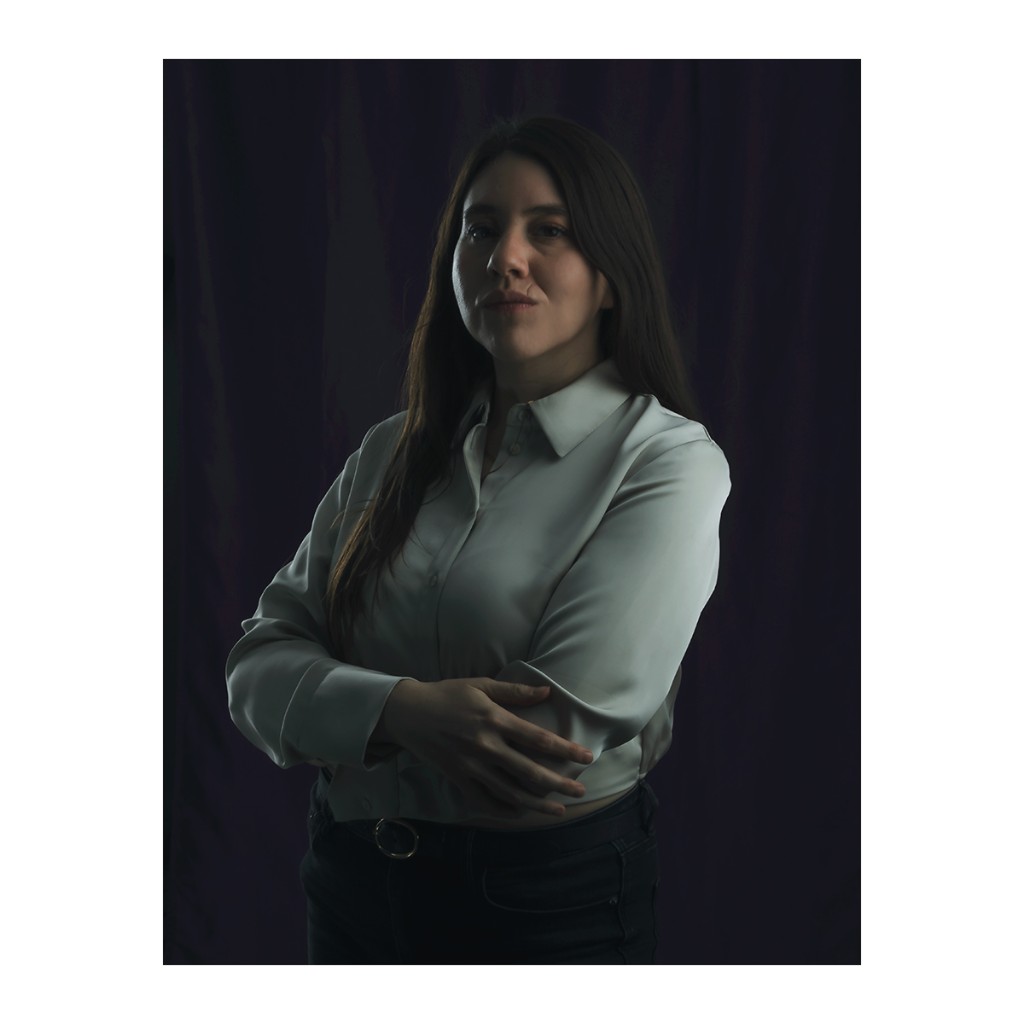 Liz Stryke authority portrait: sage green satin blouse, arms crossed with O-ring belt and dark jeans; serious direct gaze against blue-purple curtain — brand talent portrait.