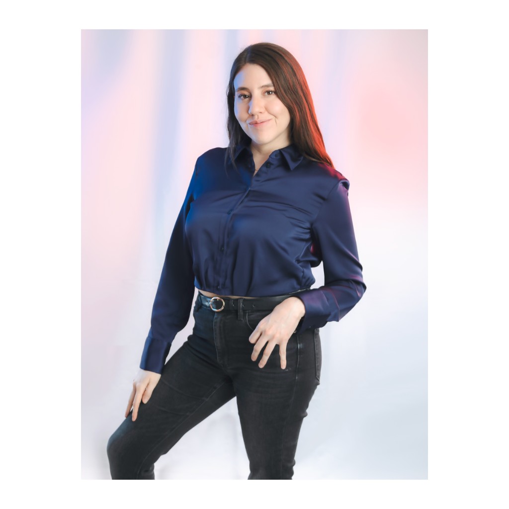 Liz Stryke lifestyle portrait: navy blue satin shirt and dark jeans, hand on hip and smile; orange-red accent light in hair on pastel gradient background — dynamic talent headshot.