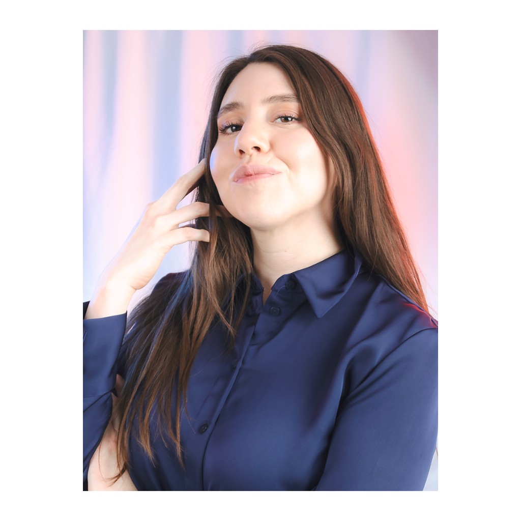 Liz Stryke color close-up: navy blue satin shirt, hand at cheek and flirtatious expression; orange-red accent light in hair; lash makeup on pastel background — editorial talent photography.
