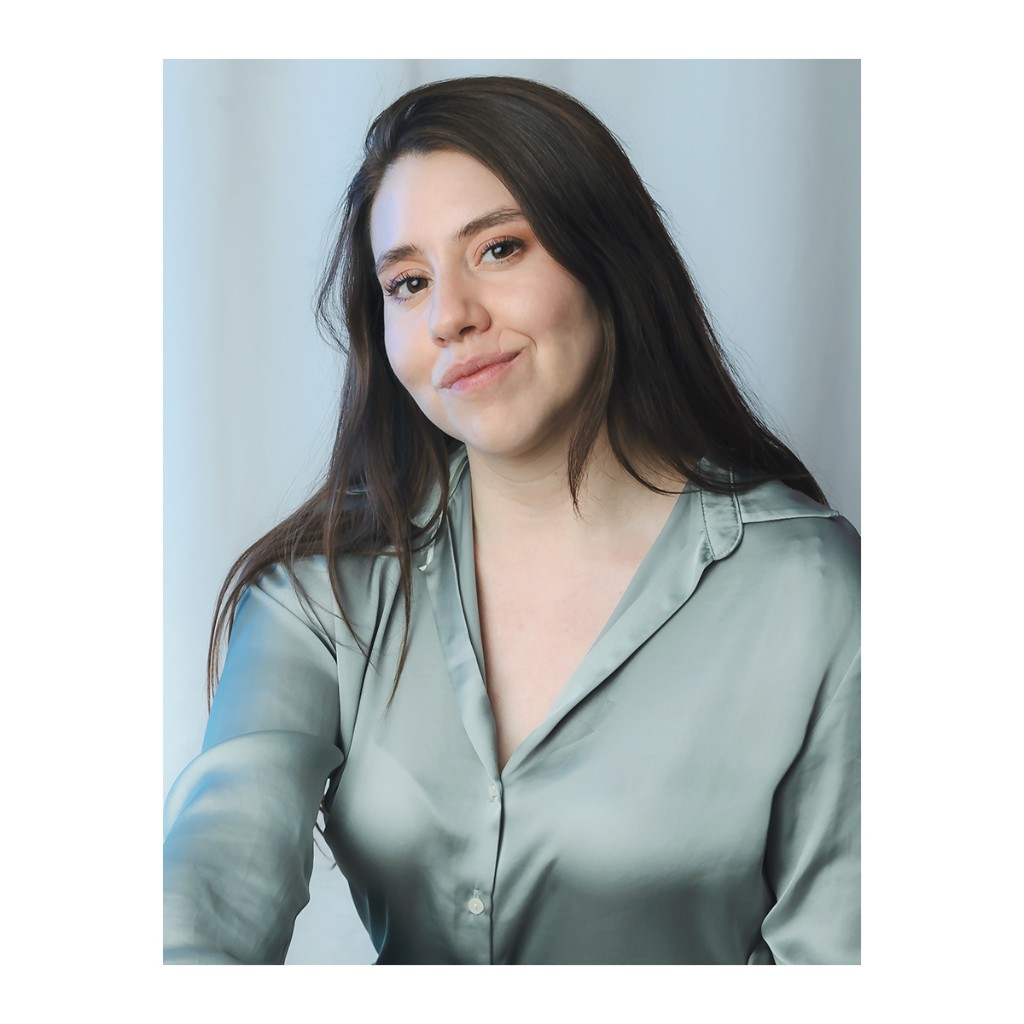 Liz Stryke close-up portrait: seafoam green satin blouse with soft open neckline, genuine smile and loose dark hair; soft light on pale blue-white backdrop — natural headshot photography.
