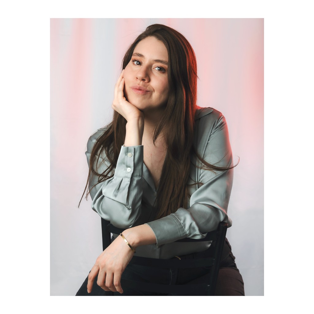 Liz Stryke color studio portrait: sage green satin blouse, chin resting on hand and warm expression; gold bangle and orange rim light in hair on pastel backdrop — editorial talent session.