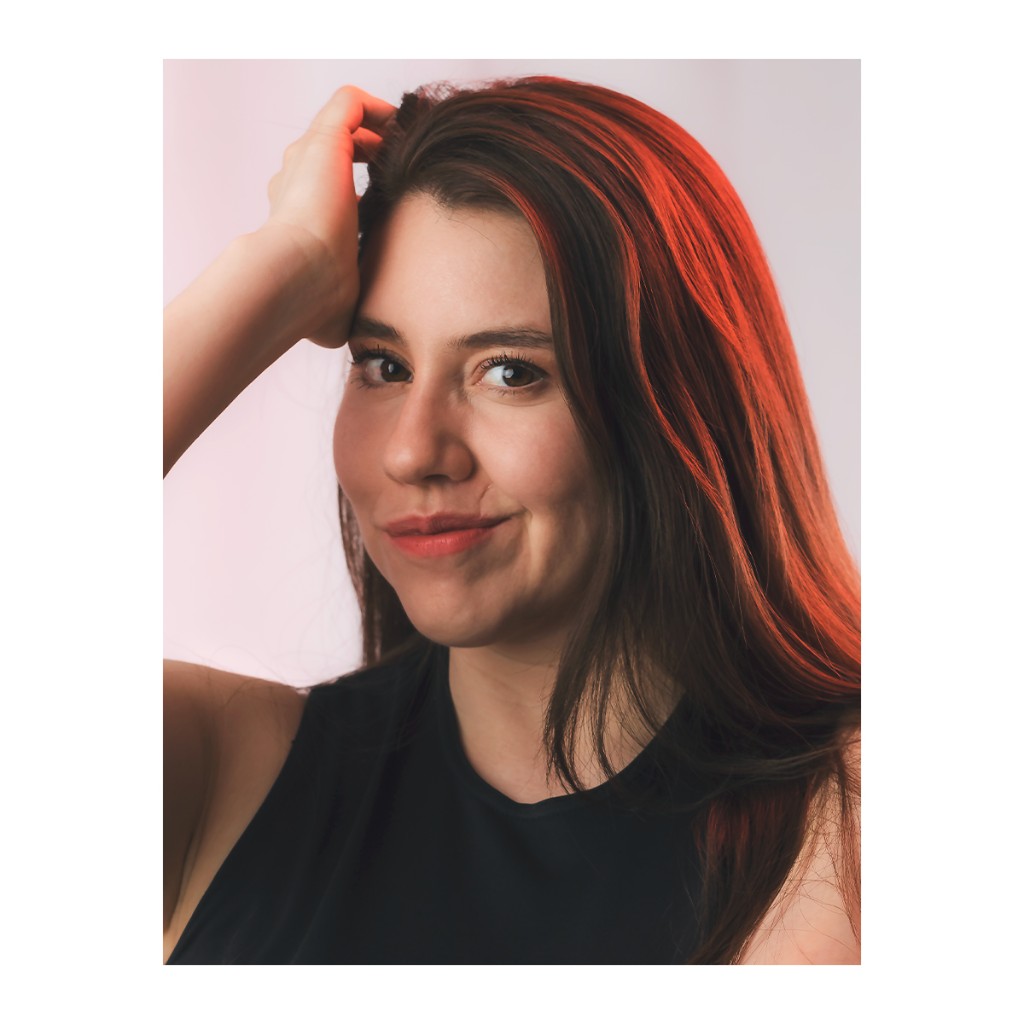 Liz Stryke editorial headshot: black racerback tank, both hands raised at the crown of the hair and relaxed smile; intense red-orange light bathing the hair on pale white background — expressive talent portrait with dramatic color.