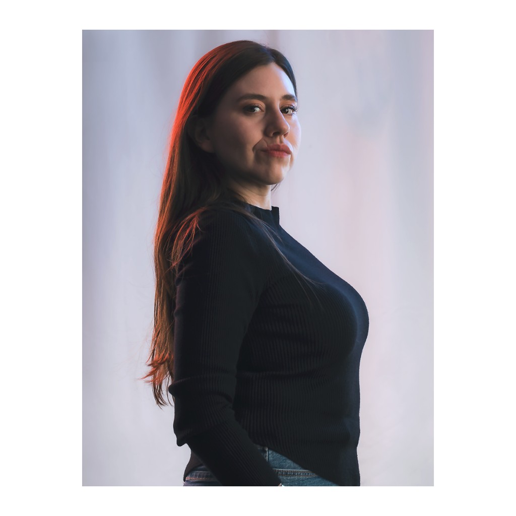 Liz Stryke profile portrait: black ribbed mock-neck long-sleeve top, dark jeans; bright orange-red rim light in hair; serene confident expression on light background — color talent headshot.