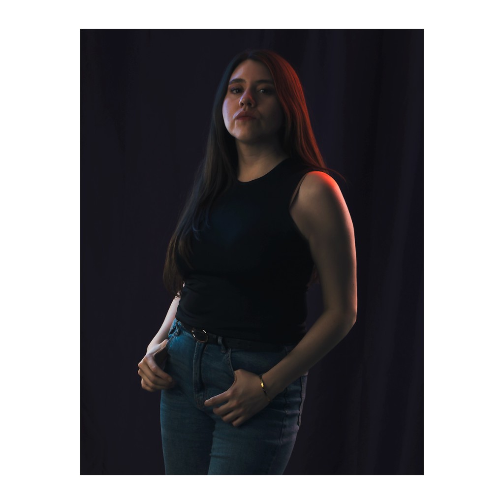 Liz Stryke full-length portrait: black sleeveless ribbed tank, light-wash blue jeans and ring-buckle belt, hands at waistband; orange-red rim light on shoulder against dark indigo curtain — editorial talent photography.
