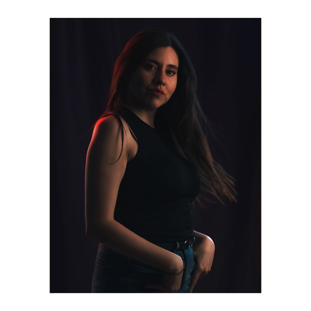 Liz Stryke portrait in motion: black racerback tank, dark jeans and ring belt, hip turned and hair in motion; orange-red rim light on arm against dark curtain — dynamic character talent session.