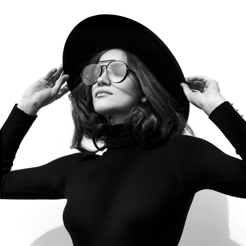Naty Velázquez B&W portrait: turtleneck, wide-brim hat and aviator glasses, hands on brim, directional light on white studio backdrop.