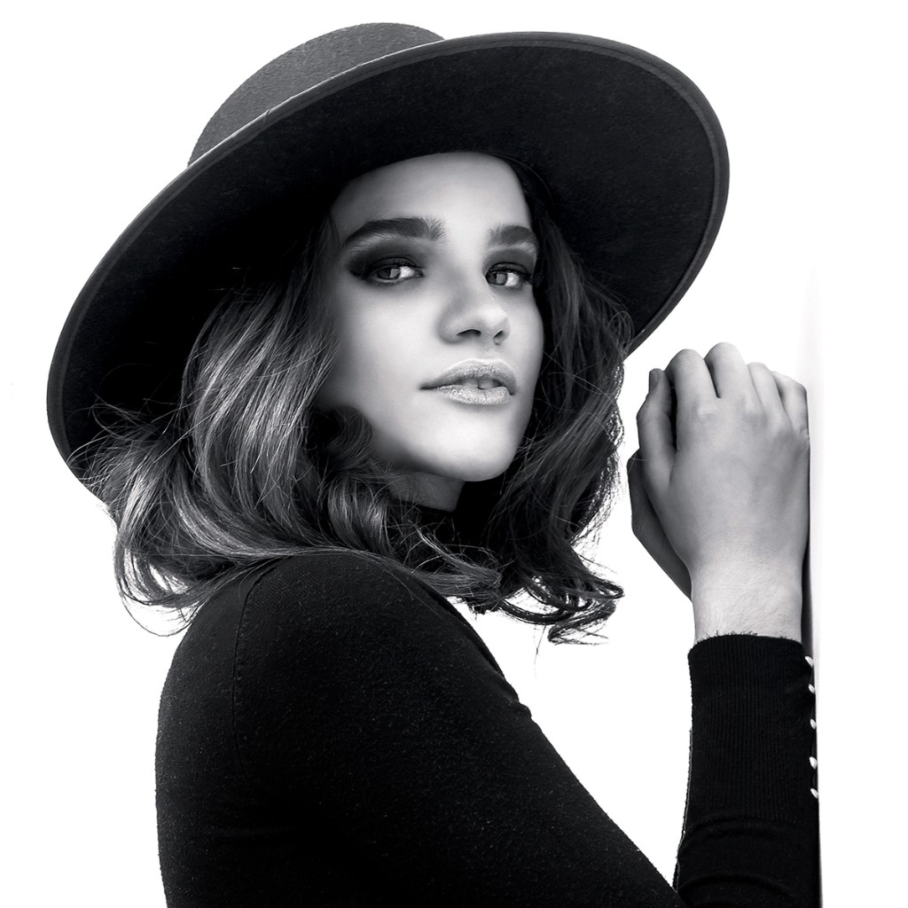 Naty Velázquez B&W portrait: wide-brim hat and black top, looking over shoulder, hands on white vertical surface.