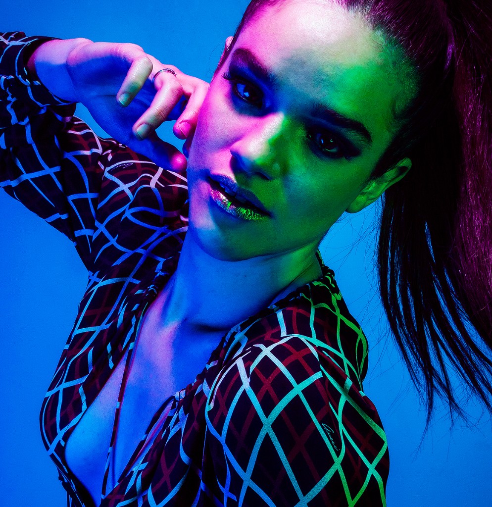 Naty Velázquez editorial close-up: sleek ponytail, smoky makeup, blue, magenta and green gels on grid-pattern top.