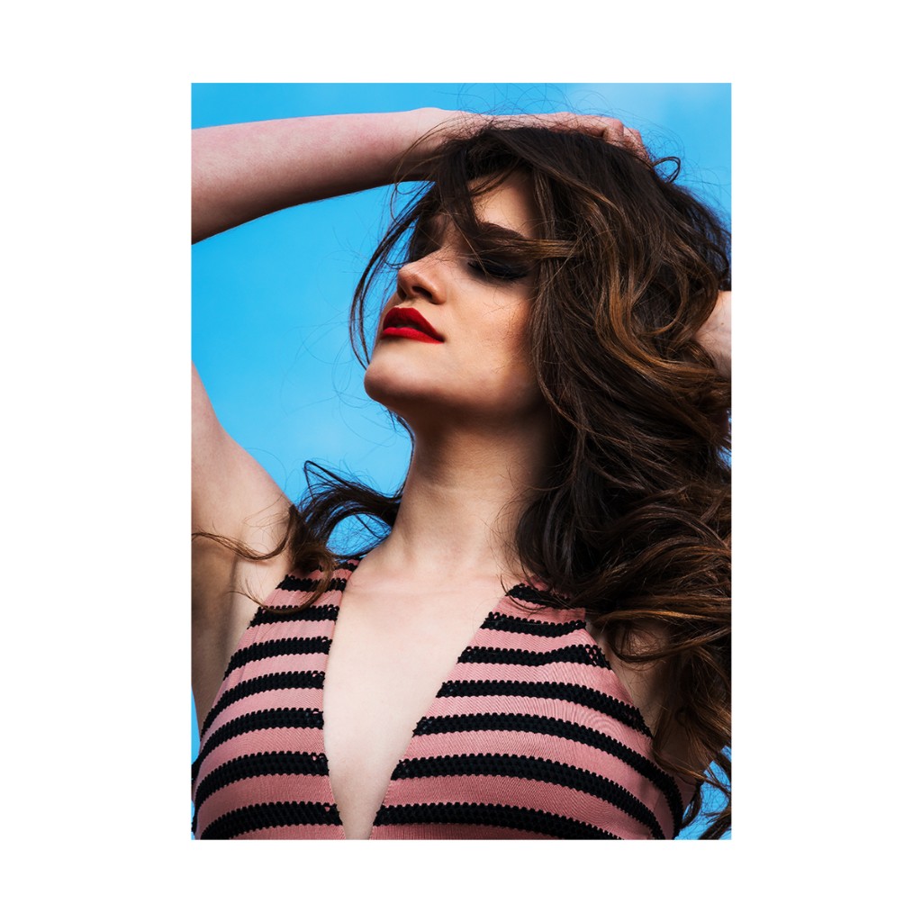 Naty Velázquez fashion portrait: hands in long wavy hair, striped deep-V top, clear blue sky in daylight.